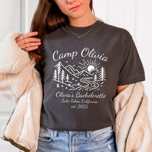 Op de afbeelding: Donkergrijs t-shirt met een wit grafisch ontwerp. Het ontwerp bevat de tekst "Camp Olivia" boven een berg- en rivierlandschap, en de tekst "Olivia's Bachelorette Lake Tahoe, California est. 2025".