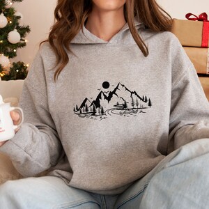 K&ouml;nnte beinhalten: Ein grauer Kapuzenpullover mit einem schwarzen Bergmotiv auf der Vorderseite. Das Motiv zeigt eine Bergkette, eine Sonne, einen Fluss und B&auml;ume.