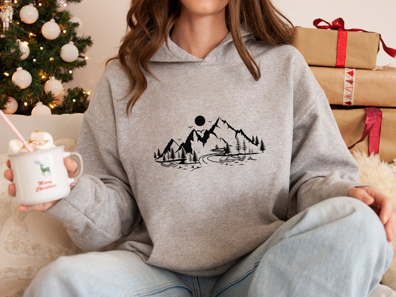 K&ouml;nnte beinhalten: Ein grauer Kapuzenpullover mit einem schwarzen Bergmotiv auf der Vorderseite. Das Motiv zeigt eine Bergkette, eine Sonne, einen Fluss und B&auml;ume.