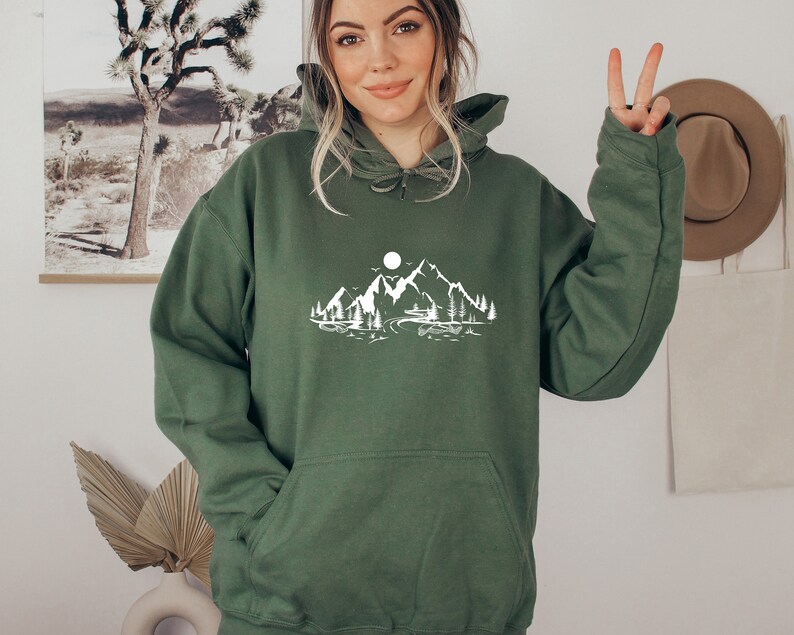 K&ouml;nnte beinhalten: Ein gr&uuml;ner Kapuzenpullover mit einem wei&szlig;en Aufdruck von Bergen, einem See und einem Mond.