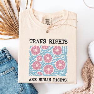 May include: A beige t-shirt with a floral design and the text "TRANS RIGHTS ARE HUMAN RIGHTS".