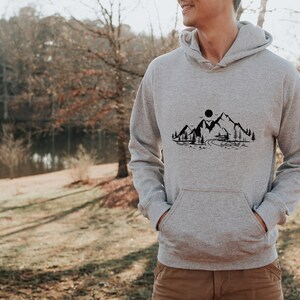 K&ouml;nnte beinhalten: Ein grauer Kapuzenpullover mit einem schwarzen Bergpanorama-Design auf der Vorderseite. Das Design beinhaltet eine kleine H&uuml;tte, einen Fluss und eine Sonne.