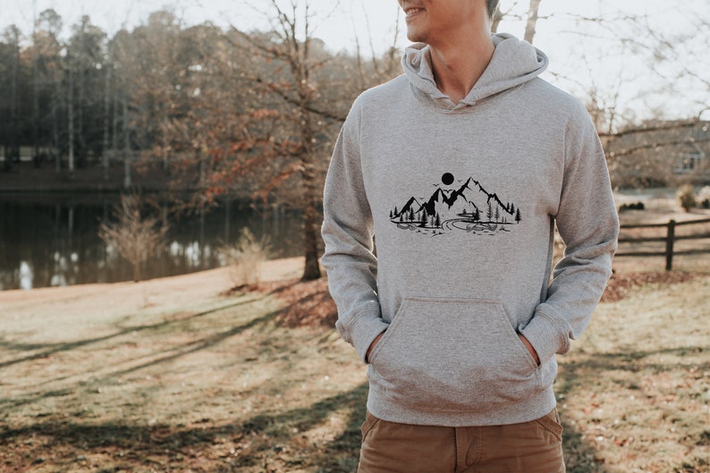 K&ouml;nnte beinhalten: Ein grauer Kapuzenpullover mit einem schwarzen Bergpanorama-Design auf der Vorderseite. Das Design beinhaltet eine kleine H&uuml;tte, einen Fluss und eine Sonne.