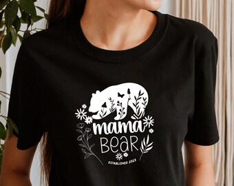 May include: A black t-shirt with a white graphic of a mama bear with flowers and butterflies. The text "mama bear established 2023" is printed on the shirt.