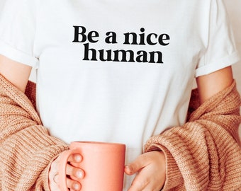 May include: A white t-shirt with the text "Be a nice human" printed in black. The person is wearing a brown cardigan and holding a light pink mug.