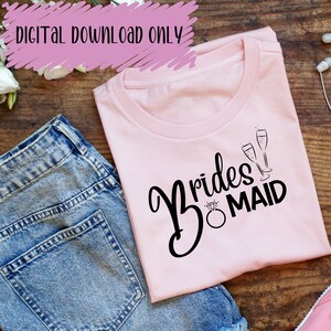 Op de afbeelding: Een roze T-shirt met de tekst "Bridesmaid" in zwarte schrijfletters met een diamanten ring en twee champagne glazen.