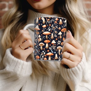 May include: White ceramic mug featuring a dark blue background with a pattern of orange and white mushrooms, small orange flowers, and white sprigs. The mug has a white handle and is held by a person wearing a white sweater.
