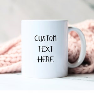 Può includere: Tazza da caffè in ceramica bianca con testo nero che recita "CUSTOM TEXT HERE".