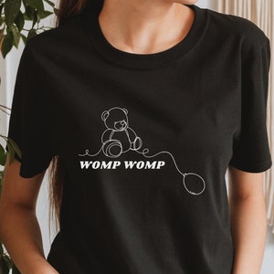 Puede incluir: Camiseta negra con un dibujo de línea blanca de un osito de peluche sosteniendo un globo. El texto "WOMP WOMP" está impreso debajo del oso.