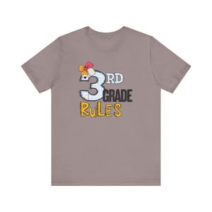 May include: A short-sleeved, taupe t-shirt featuring the text "3rd Grade Rules" in a playful design. The number "3" is blue with white stripes, and the word "Rules" is in yellow. A colorful bow adorns the top left of the number.