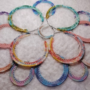 May include: A collection of colorful, crocheted hoop earrings. The earrings feature a variety of colors, including pink, blue, yellow, and green. The hoops are arranged in a circular pattern, creating a visually appealing display. The earrings are likely handmade.