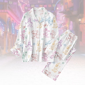 Op de afbeelding: Witte pyjamaset met kleurrijke omtrekken van stripfiguren. De top met lange mouwen heeft een knoopsluiting en een reverskraag. De bijpassende broek heeft een elastische tailleband. De set is versierd met sterren en strikken.