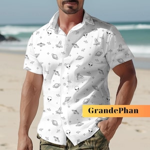 Könnte beinhalten: Ein weißes, kurzärmeliges Hemd mit Knöpfen und einem sich wiederholenden Muster aus schwarzen Alienköpfen, Raumschiffen und Planeten. Das Hemd hat einen Kragen und eine Knopfleiste vorne. Der Text "GrandePhan" ist in der rechten unteren Ecke des Hemdes aufgedruckt.