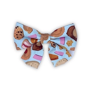 May include: A light blue bow with a pattern of various cookies and pastries. The bow is made of fabric and has a slightly textured surface.