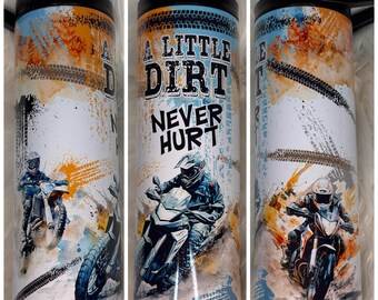 May include: A white tumbler with a black lid and handle, featuring a design with dirt bike riders and tire tracks. The text "A Little Dirt Never Hurt" is prominently displayed, with orange, blue, and brown accents.