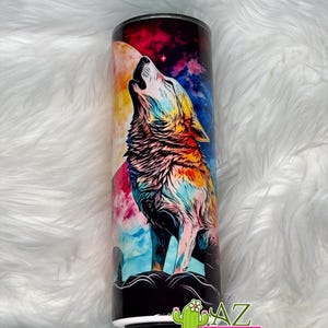 May include: A stainless steel tumbler featuring a colorful illustration of a howling wolf against a backdrop of a full moon and a starry night sky. The wolf is depicted in vibrant hues, with a mix of blues, oranges, and yellows. The tumbler has a black rim and base.