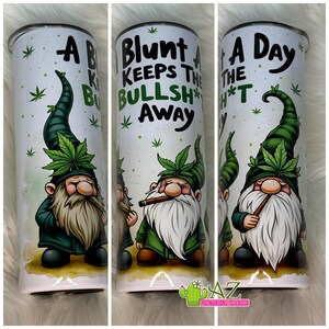 May include: A white cylindrical tumbler featuring a cartoon gnome with a long white beard, a green hat adorned with a cannabis leaf, and a brown cigar. The text reads A Blunt A Day Keeps The Bullsh*t Away, surrounded by small green cannabis leaf icons.