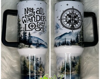 May include: A white stainless steel travel mug with a black handle and lid. The mug features a watercolor design of a mountain range with a compass and the text "Not all who wander are lost".