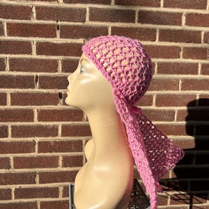 May include: A pink crocheted head covering with a net-like design and a long, flowing tail. The head covering is displayed on a mannequin head against a brick wall background. The head covering is a light pink color.