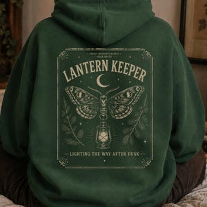 May include: A forest green hooded sweatshirt featuring a graphic on the back. The design includes a moth above a lantern, surrounded by pine branches and a crescent moon. The text reads LANTERN KEEPER and LIGHTING THE WAY AFTER DUSK.