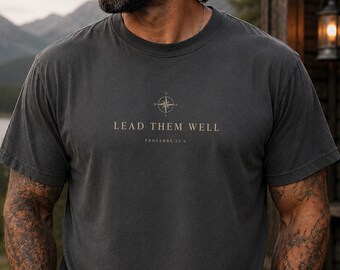 May include: A charcoal gray short-sleeve crew neck t-shirt featuring a compass graphic centered on the chest above the text "LEAD THEM WELL" and "PROVERBS 22:6" printed in a light beige color. The shirt has a standard fit and plain hem.