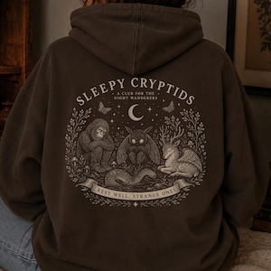 May include: A brown hoodie with the text "SLEEPY CRYPTIDS" and an illustration of cryptids. The design includes a Bigfoot, a Mothman, and a deer-like creature, with the words "REST WELL, STRANGE ONES" on a banner below. The hoodie is worn by a person.