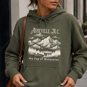 May include: Military green hoodie with a front pocket and drawstring. The hoodie features a white graphic of mountains and the text "Asheville, N.C." and "On Top of Mountains". The bottom of the hoodie is labeled "military green".