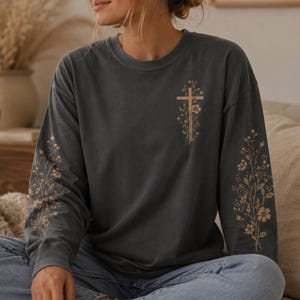 May include: A charcoal gray long-sleeve crewneck sweatshirt featuring a light tan graphic of a cross intertwined with delicate floral vines on the left chest and matching floral vine designs printed along both sleeves.