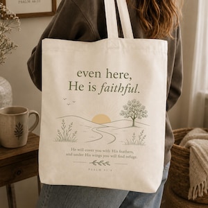 Op de afbeelding: Een crèmekleurige canvas draagtas met een gedrukte landschapsillustratie van een zon, heuvels en een boom. De tekst luidt: even here, He is faithful. He will cover you with His feathers, and under His wings you will find refuge. Psalm 91:4.