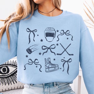 May include: Light blue sweatshirt with a graphic design of hockey equipment and bows. The design is in a distressed style and includes a hockey helmet, hockey stick, hockey puck, and ice skates.
