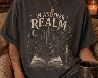 May include: A charcoal gray short-sleeve t-shirt featuring a graphic print of an open book with a castle, roses, a raven, and a candle. The text "IN ANOTHER REALM" is printed in a decorative font above the illustration, which also includes stars and a crescent moon.