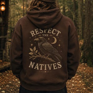 May include: A dark brown hooded sweatshirt featuring a back graphic of a raven perched on a branch. The design includes a crescent moon, stars, and the text "RESPECT THE NATIVES" in a light beige font. The hoodie has long sleeves and a hood.