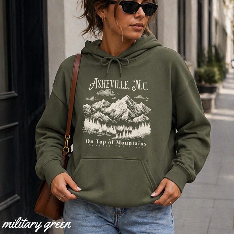 May include: Military green hoodie with a front pocket and drawstring. The hoodie features a white graphic of mountains and the text "Asheville, N.C." and "On Top of Mountains". The bottom of the hoodie is labeled "military green".