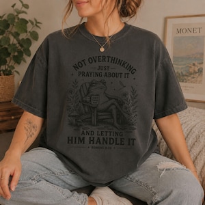 May include: A charcoal gray short-sleeve t-shirt featuring a graphic of a frog sitting in a chair holding a mug. The text reads: NOT OVERTHINKING JUST PRAYING ABOUT IT AND LETTING HIM HANDLE IT ROMANS 8:28. The design includes flowers and a mushroom.