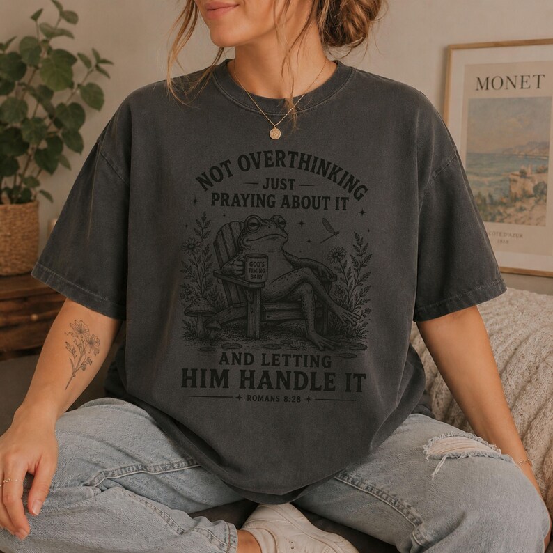 May include: A charcoal gray short-sleeve t-shirt featuring a graphic of a frog sitting in a chair holding a mug. The text reads: NOT OVERTHINKING JUST PRAYING ABOUT IT AND LETTING HIM HANDLE IT ROMANS 8:28. The design includes flowers and a mushroom.