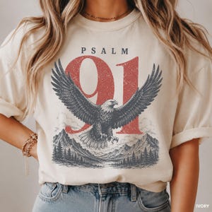 May include: An ivory crewneck t-shirt featuring a graphic print of a soaring eagle over a mountain landscape. The text PSALM 91 is printed in black above a large, distressed red number 91. The eagle and mountain scene are rendered in dark grey.