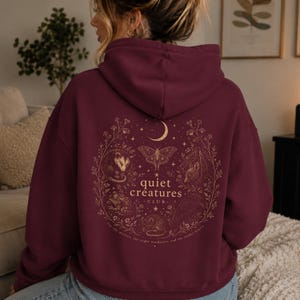 K&ouml;nnte beinhalten: Burgunderfarbener Kapuzenpullover mit goldfarbener Grafik auf dem R&uuml;cken. Das Design zeigt einen Mond, Sterne, eine Motte und verschiedene Tiere in einem floralen Kranz. Der Text lautet "quiet creatures club" und "for the misfits, the night wanderers, and the tender hearts."