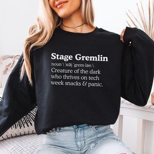 May include: Black sweatshirt with the text "Stage Gremlin" and a definition. The definition describes a creature thriving on tech, snacks, and panic. The sweatshirt is a crew neck style.