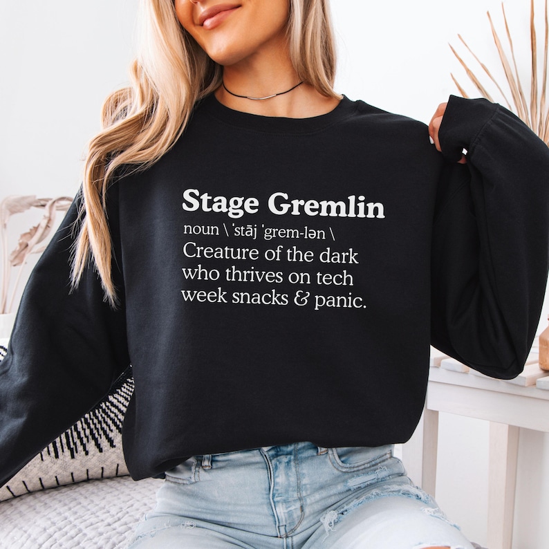 May include: Black sweatshirt with the text "Stage Gremlin" and a definition. The definition describes a creature thriving on tech, snacks, and panic. The sweatshirt is a crew neck style.