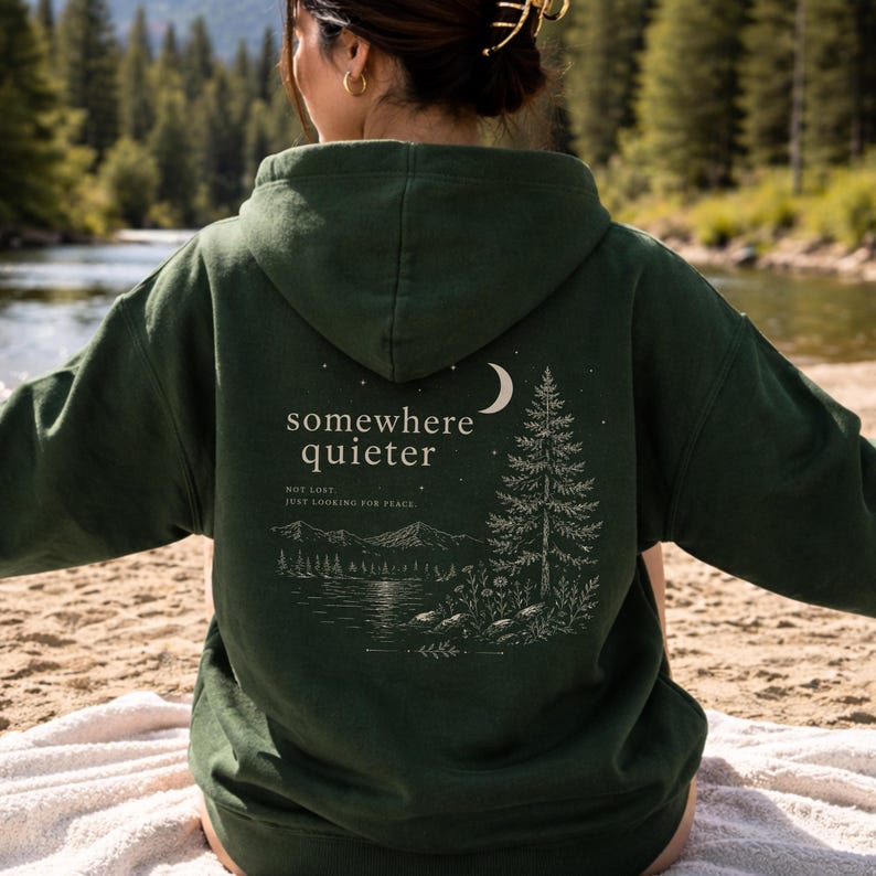 May include: Dark green hoodie with a hood and a graphic on the back. The graphic features a crescent moon, a pine tree, and the words "somewhere quieter" above a landscape scene. The text "not lost, just looking for peace" is below the main text.