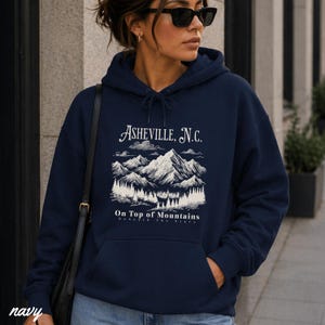 May include: Navy blue hoodie with a drawstring hood and a front pocket. The front of the hoodie features a white graphic of mountains, trees, and the text "Asheville, N.C." and "On Top of Mountains". The word "navy" is printed at the bottom left.