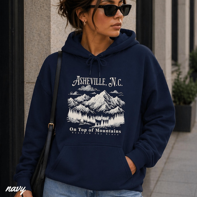 May include: Navy blue hoodie with a drawstring hood and a front pocket. The front of the hoodie features a white graphic of mountains, trees, and the text "Asheville, N.C." and "On Top of Mountains". The word "navy" is printed at the bottom left.