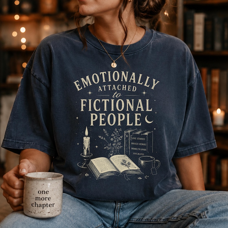 May include: Navy blue t-shirt with the text "Emotionally Attached to Fictional People." The design includes a candle, open book, books, and a mug that says "one more chapter." A cosy, book-themed graphic tee.