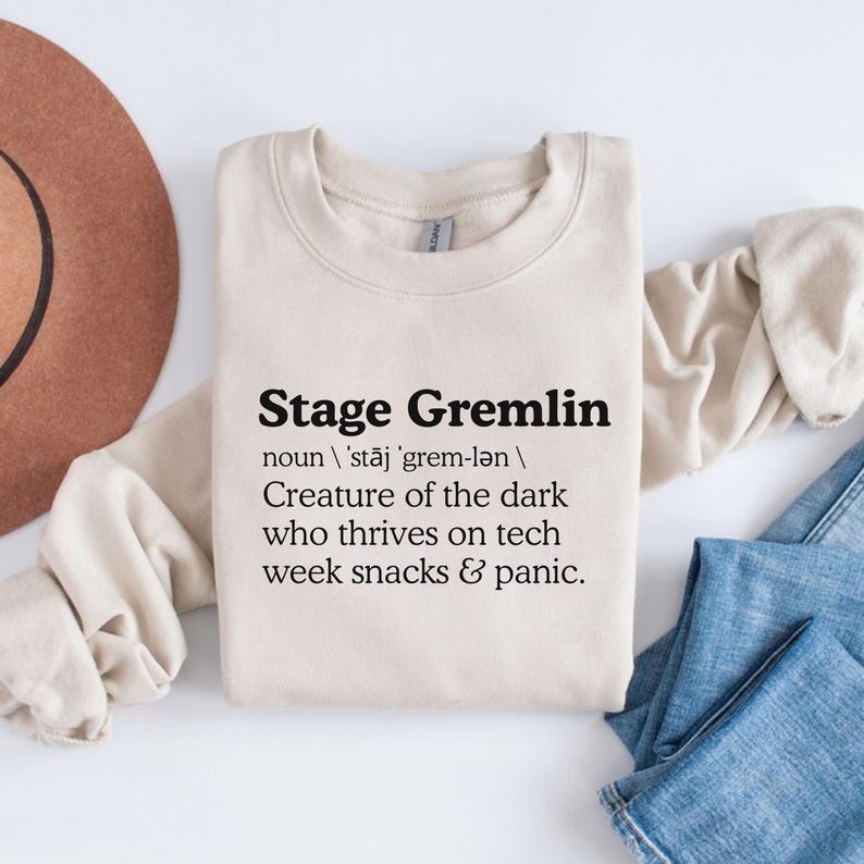 May include: A cream-colored sweatshirt with the text "Stage Gremlin" in bold black letters. The definition below describes a creature thriving on tech week snacks and panic. A brown hat and blue jeans are in the background.