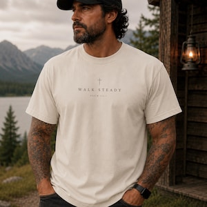 K&ouml;nnte beinhalten: Ein cremefarbenes Kurzarm-T-Shirt mit einem kleinen schwarzen Kreuz-Symbol, das mittig &uuml;ber dem Text "WALK STEADY" und "PSALM 112:7" in einer schlichten, klaren Schrift auf der Brust des Kleidungsst&uuml;cks gedruckt ist.