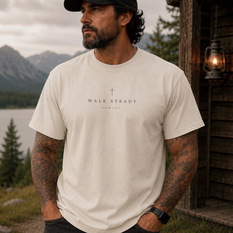 K&ouml;nnte beinhalten: Ein cremefarbenes Kurzarm-T-Shirt mit einem kleinen schwarzen Kreuz-Symbol, das mittig &uuml;ber dem Text "WALK STEADY" und "PSALM 112:7" in einer schlichten, klaren Schrift auf der Brust des Kleidungsst&uuml;cks gedruckt ist.