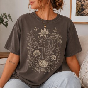 May include: A charcoal gray short-sleeve t-shirt featuring a light-colored line art graphic on the front. The design displays a central cross surrounded by various wildflowers, foliage, and two small butterflies arranged in a vertical botanical pattern.