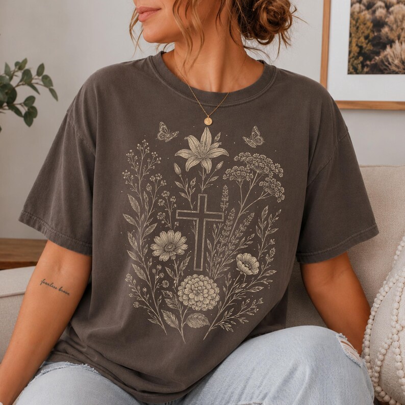May include: A charcoal gray short-sleeve t-shirt featuring a light-colored line art graphic on the front. The design displays a central cross surrounded by various wildflowers, foliage, and two small butterflies arranged in a vertical botanical pattern.