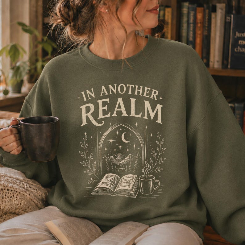 Może przedstawiać: Oliwkowa bluza z okrągłym dekoltem i kremowym nadrukiem graficznym. Napis "IN ANOTHER REALM" znajduje się nad ilustracją przedstawiającą otwartą książkę, parujący kubek oraz łukowate okno z g&oacute;rskim krajobrazem pod p&oacute;łksiężycem.