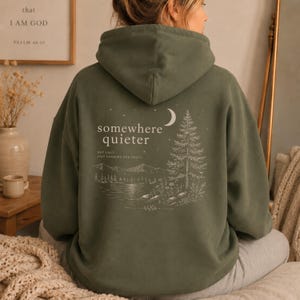 May include: Olive green hoodie with a white graphic on the back featuring a crescent moon, pine tree, lake, and the words "somewhere quieter." The text below reads "not lost just looking for peace." The hoodie has a drawstring hood.